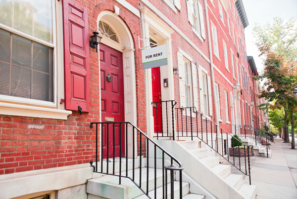 A row of houses, with a sign saying “For rent” hanging by the door of one of the houses.