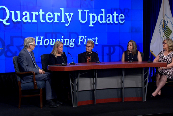 Panelists sit at a table in front of a backdrop that says “Quarterly Update Housing First”.