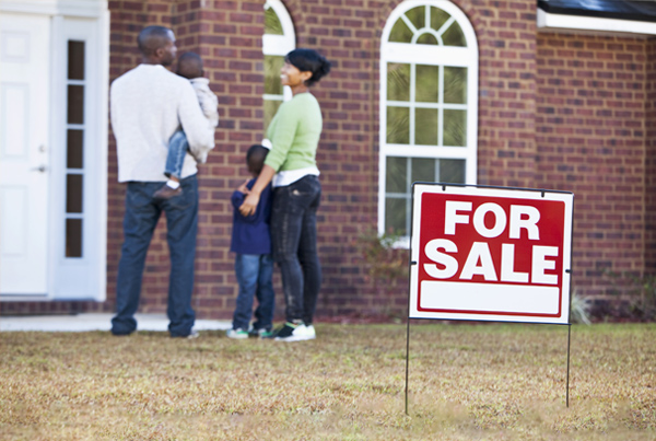 A family in front of a house with a “For Sale” sign.
