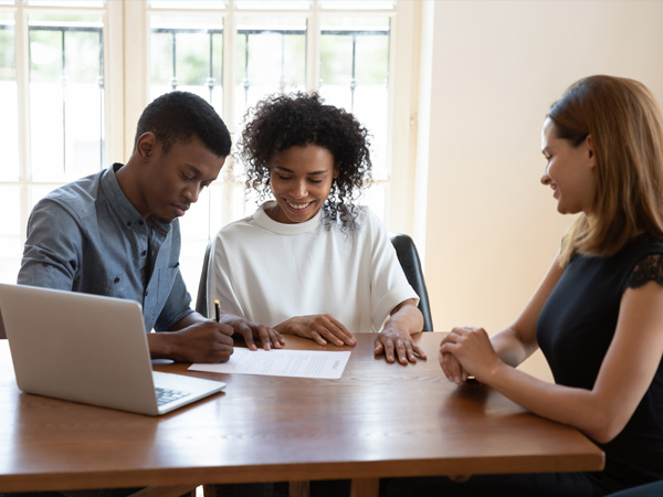 Photo of a couple sitting at a table and signing a document and a realtor sitting next to them.