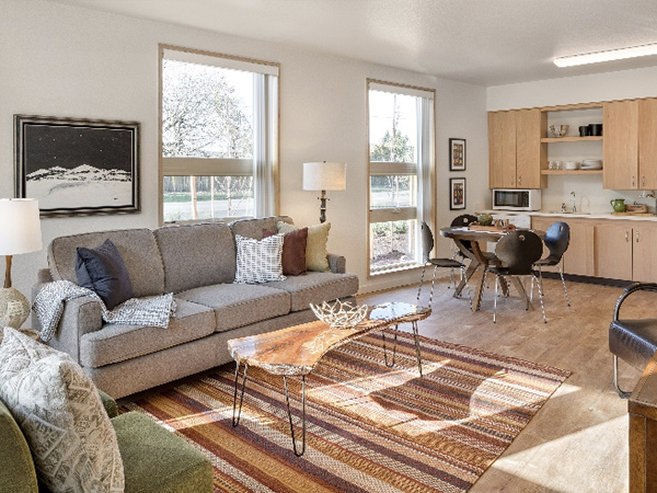 Photograph of living room that includes a three-seater couch, a rug, and coffee table with a dining table and chairs situated in front of a kitchen unit along the back wall.