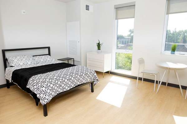 An interior unit with a bed, a side desk, a dresser, and table with a chair in front of a window.