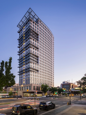 A corner lot housing a 26-story building with a street and parked cars in the foreground.