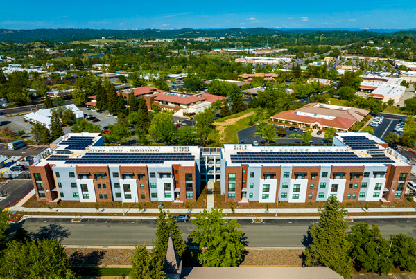 Low-angle aerial photograph of the street-facing facade of two three-story buildings with buildings, trees, and mountains in the background.