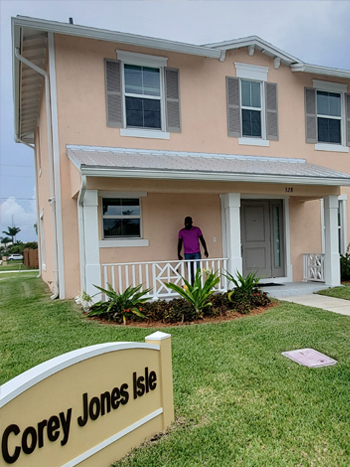 Photograph of a smiling man standing on the front porch of a two-story peach-colored house, with a sign that reads “Corey Jones Isle” on the front lawn.