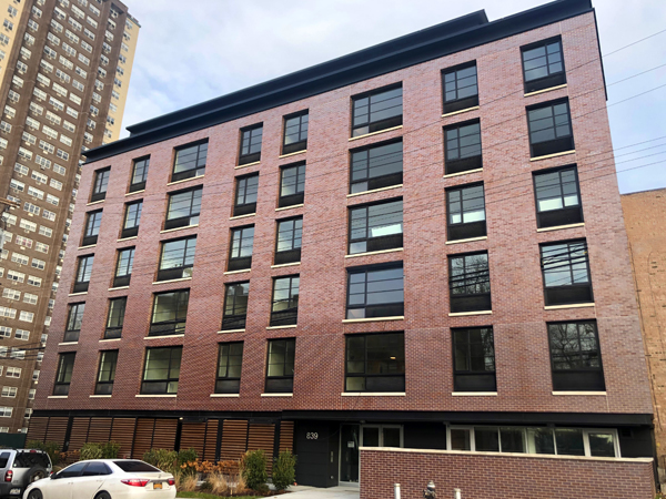 Photograph of the front façade of a seven-story red brick residential building with a sixth-floor setback, flanked by other brick buildings. 