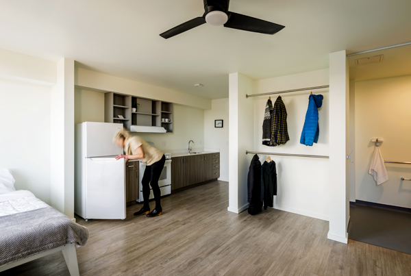 Studio apartment including a bed and a kitchen with a woman getting something from the refrigerator. Studio apartment including a bed and a kitchen with a woman getting something from the refrigerator.