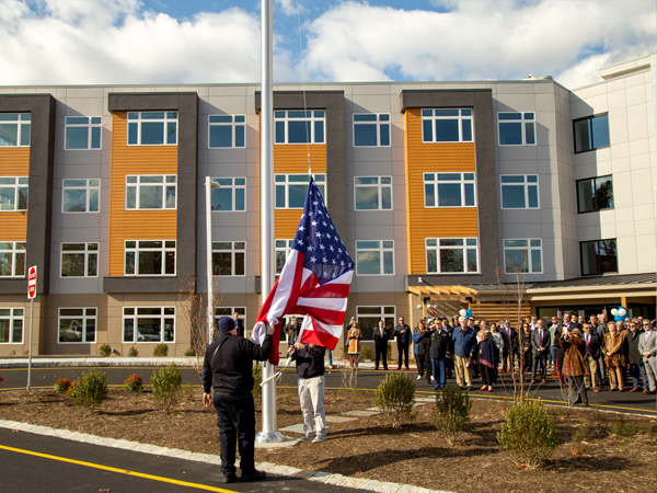 Photograph of flag raising ceremony with a residential building in the background. 