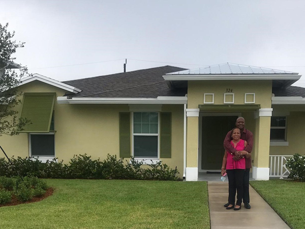 Photograph of a smiling couple standing in front of a one-story house painted pastel yellow, with a front porch and olive-green shutters.