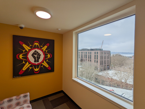 Photo of the corner of a room with a large window on the right and native artwork on the left. 