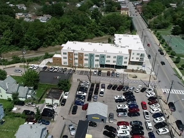 Aerial photograph of an L-shaped apartment building, bounded on two sides by streets and on one side by train tracks; across the larger street, there is a wooded recreation area with paths and a tennis court.