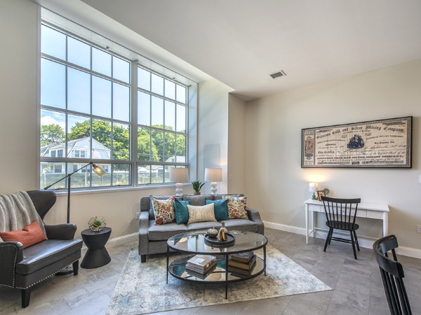 Photograph of unit interior living room containing a large window, a 3-seater couch, a chair, and a coffee table. 