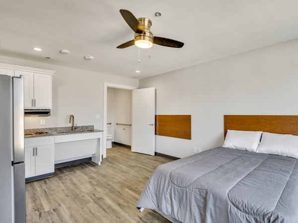 Photograph of an interior unit with a bed and refrigerator in the foreground, kitchenette along the back wall, and a door leading to a bathroom.