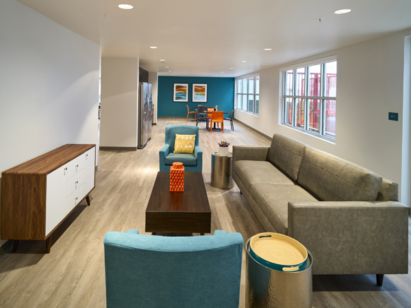 Photograph of an interior seating area containing two chairs, a coffee table, two side tables, and a sofa that is facing an ottoman along the wall with additional seating and two refrigerators in the background.  