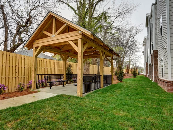 A gazebo bordered by a fence and trees on the left and grass area and the apartment building façade on the right.