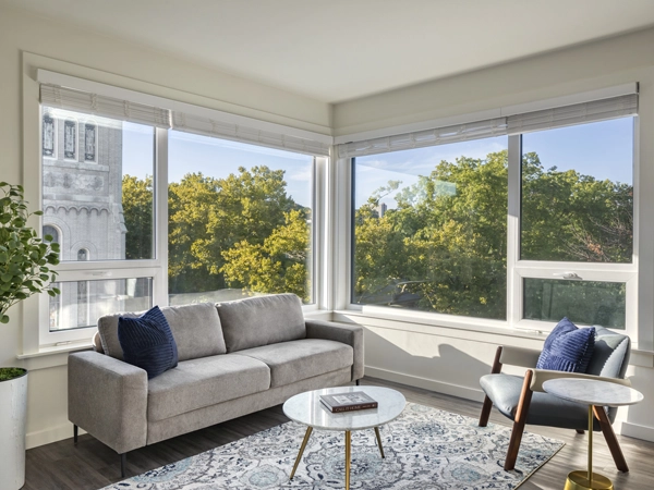 A modern living room with large corner windows, a sofa with a potted plant next to it, chair, small round tables, and a patterned rug.