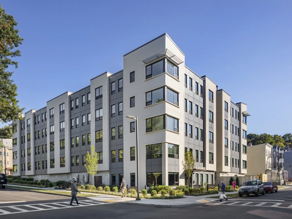 A five-story, modern gray-and-white apartment building on an urban block with crosswalks and trees nearby.