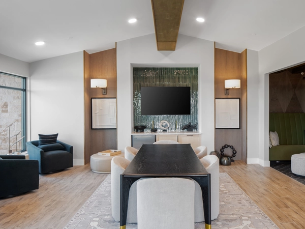 A lounge with a long dining table surrounded by chairs, with a wall-mounted television and a seating nook in the background.