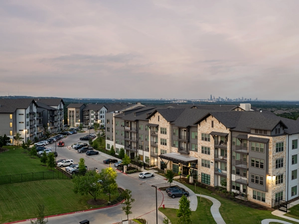 An aerial photograph of an apartment complex of multiple four-story buildings connected by a road lined with parked cars, surrounded by green lawns and a city skyline in the background. 