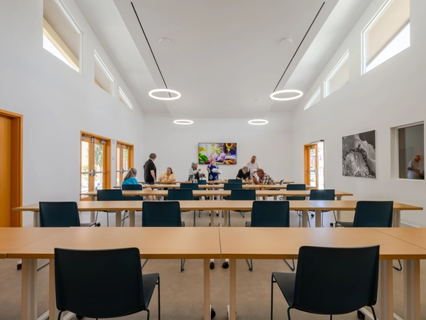 A large room with tables and chairs and people congregated at a table at the far end of the room.