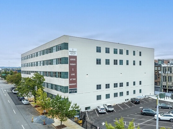 A white, five-story concrete building on a tree-lined street. A banner advertising "Market Lofts" is visible on the side of the building, which abuts a surface parking lot.