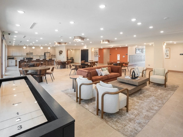 A large common room with white walls, light-colored tile flooring, and bright recessed lighting. The space includes ample seating, a kitchen area, and a gaming table.