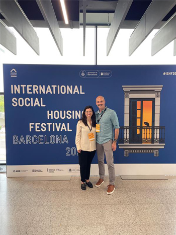 Victoria Brown (left) and Brian McCabe (right) stand behind a large poster with text advertising the International Social Housing Festival in Barcelona.