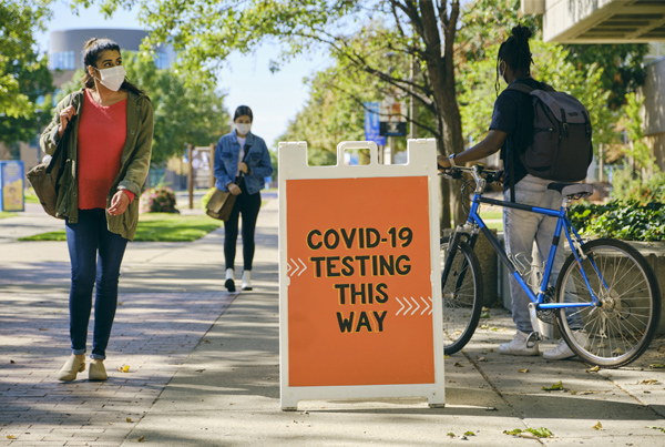 A sign on a sidewalk reads “COVID-19 testing this way” as people walk by.