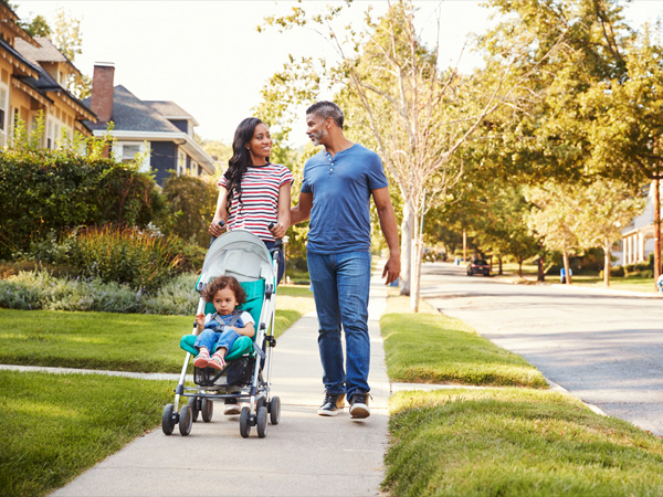 A couple walks down a sidewalk in a neighborhood while pushing a stroller with a child.