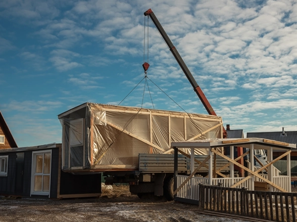 A crane hoists a house on a truck, illustrating the transportation of a building structure.