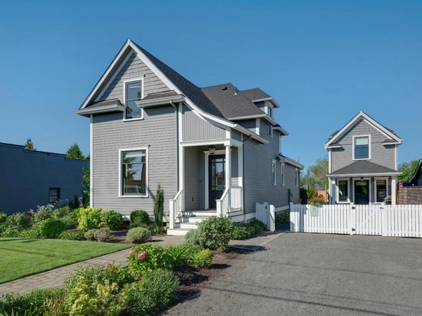 A gray house with a driveway, and a smaller house next to it and behind a white fence.