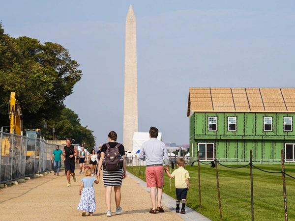 A crowd of people walking in the National Mall, with a green two-story model house on the right and the Washington Monument in the background.