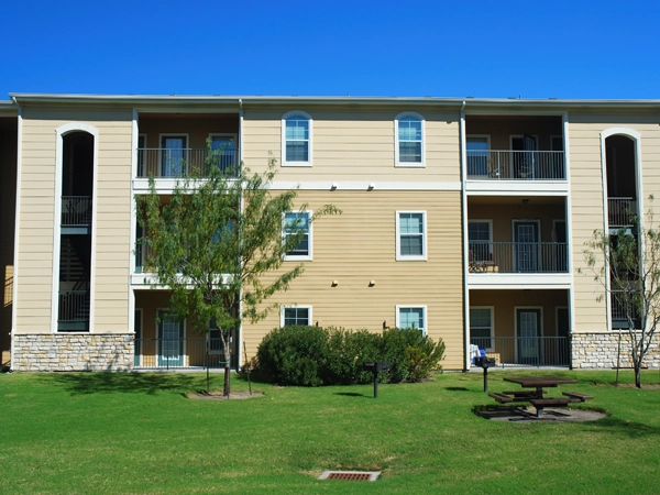 An apartment complex with a lawn, small trees and shrubbery, and a picnic table in front.