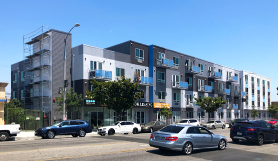 Photograph of the front and side façades of a four-story building behind a landscaped sidewalk, with a multilane street and cars in the foreground. 