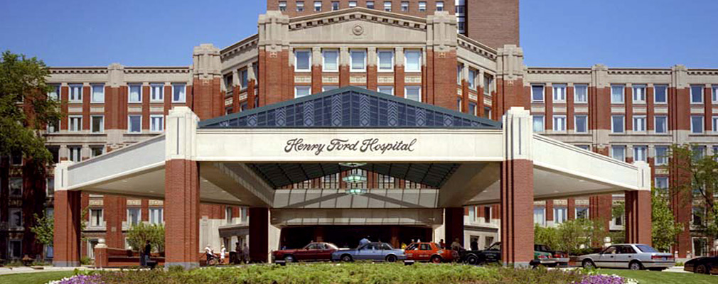 A photograph of the front façade of the Henry Ford Hospital building, a brick building six stories tall. The entrance is highlighted with a large three-sided extension, with a shallow pediment over the front side of the bay.  In front of the entrance bay is a large octagonal port cochere that has a glass pedimented roof over its center; approximately six cars are parked under the port cochere. 'Henry Ford Hospital' is written in script on the frieze of the porte cochere.