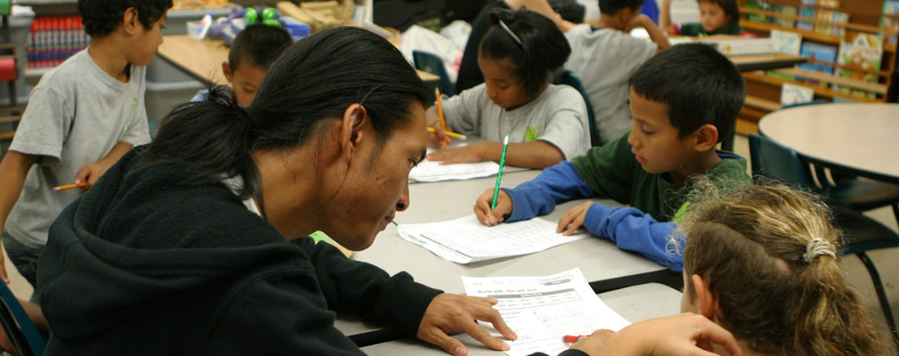 Photograph of a class room in which college students help two groups of elementary-school children with their school work.