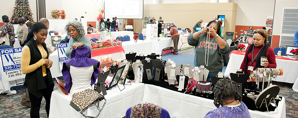 Photograph of a large meeting room set with tables displaying jewelry and other wares; groups of people talk to each other and examine the items on display. A decorated Christmas tree stands in the background.