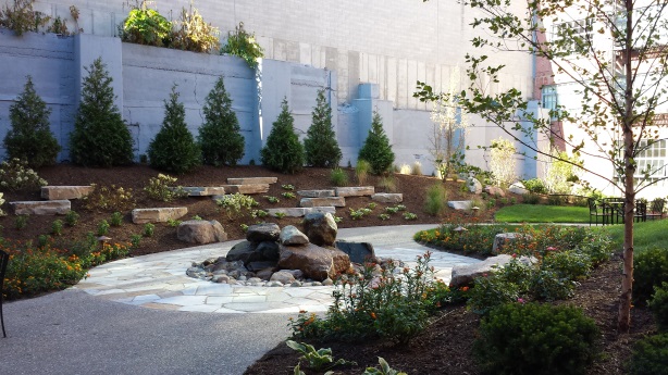 Photograph of the courtyard at Baker Lofts. An asphalt and stone walkway passes through the courtyard containing ornamental rocks, evergreen and deciduous trees, small bushes and flowering plants, and lawn. A table and chairs are at the far end of the courtyard, which is framed in the background by a masonry wall and the Baker Lofts building.
