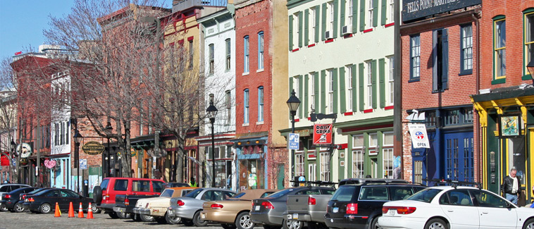 Photograph of several contiguous buildings with brightly-colored, varied façades in the historic Fell’s Point community of Baltimore, Maryland. Parked cars line the street in front of the buildings.