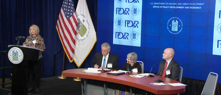 Photograph of panelist Carol Gore speaking at a podium while moderator Roger Boyd, deputy assistant secretary of HUD’s Office of Native American Programs, and panelists Kevin Klingbeil and Nancy Pindus sit at a table onstage.
