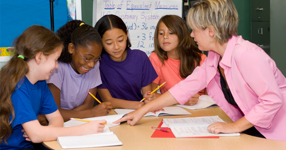 Four female students sit at a table with a female teacher (far right), writing in notebooks with pencils as the teacher points to one of the notebook pages. A chart with measurement conversions is visible in the background.