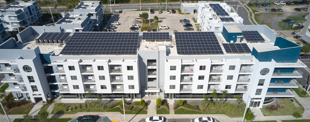 Low-angle aerial photograph of a four-story residential building, with rooftop solar panels and with two multistory buildings and a parking lot behind the larger building. 