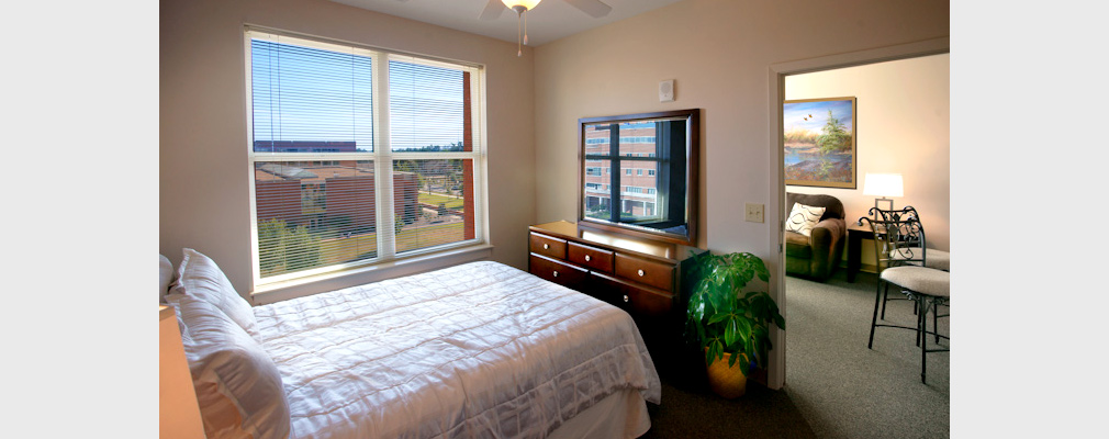 Photograph of the interior of a bedroom with a doorway opening into the living room and window overlooking a university building.