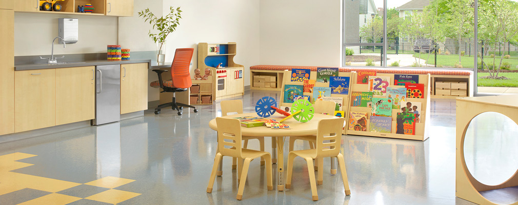 Photograph of the interior of a classroom containing a table and four chairs, a book display, and play equipment, with a landscaped play area beyond large windows in the back wall. 