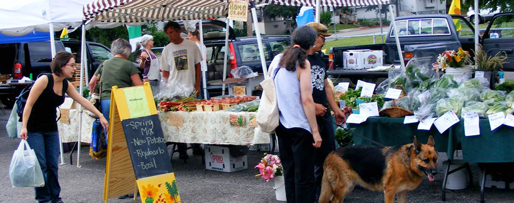 Photograph of people shopping at an outdoor market, with tables of several vegetable vendors under canopies in a parking lot.