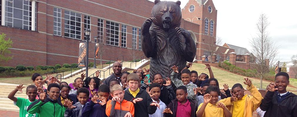 Photograph of approximately 30 elementary school students and a teacher in front of a statue of a bear, the university’s mascot.