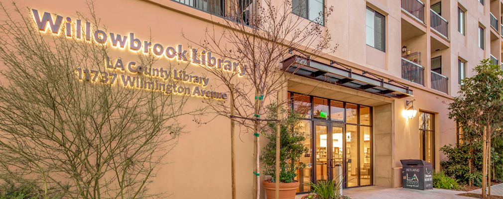 Photograph of an entrance to a multistory mixed-use building, with a wall sign near the door reading in part, “Willowbrook Library: LA County Library." 