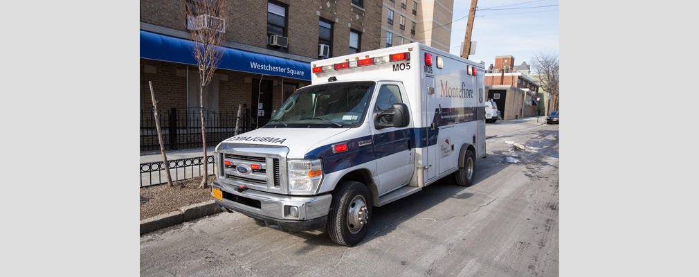 Photograph of an ambulance parked on a street, with multistory brick buildings in the background.