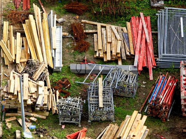Photograph of wooden planks and metal building materials arranged in piles on a vegetated lot.