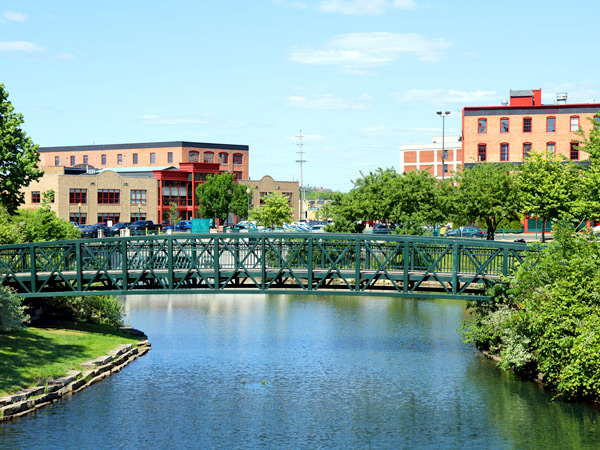 Downtown Kalamazoo, Michigan. A bridge over water is visible in the foreground, and multistory buildings and trees are visible in the background.